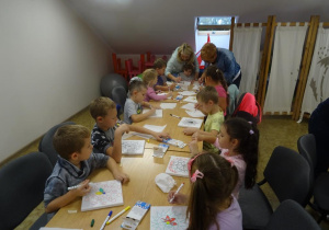 Grupa dzieci siedzi przy długim stole i maluje pisakami na płótnie. Nas tsołem pochylona jest pani dyrektor i pani Ewa, które pomagają dzieciom podczas malowania.